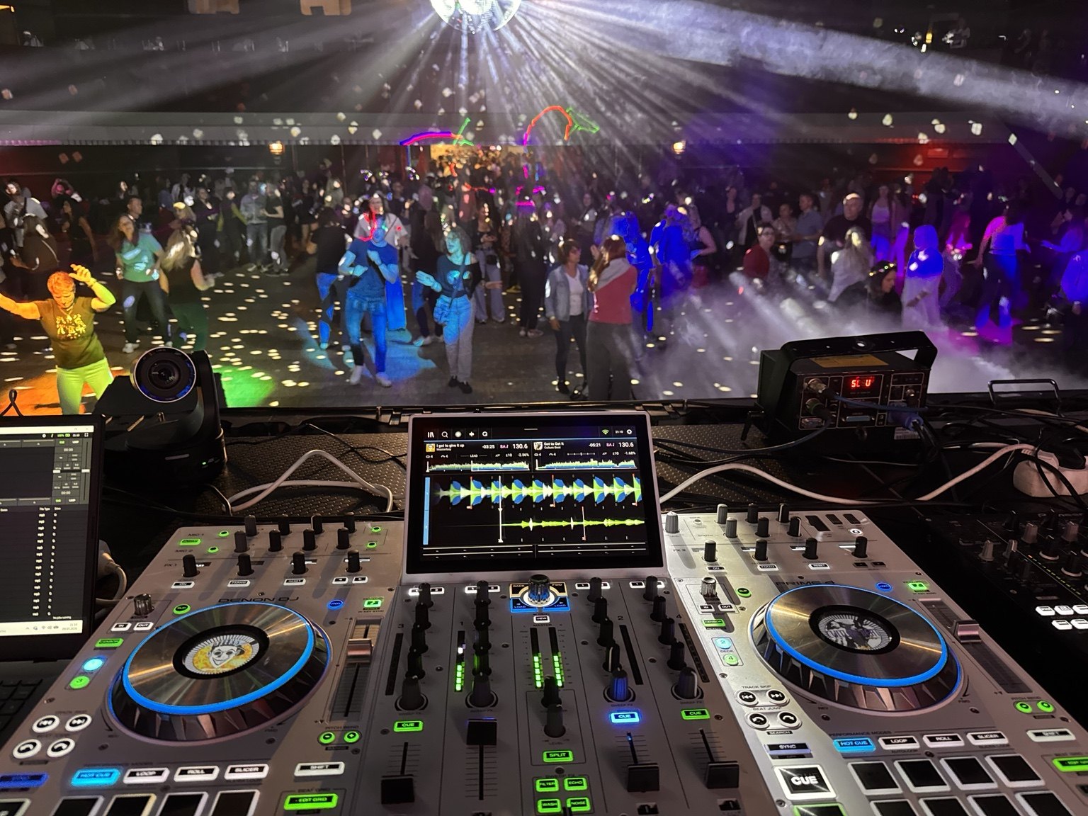 Plný taneční parket pod světly, v popředí DJ pult